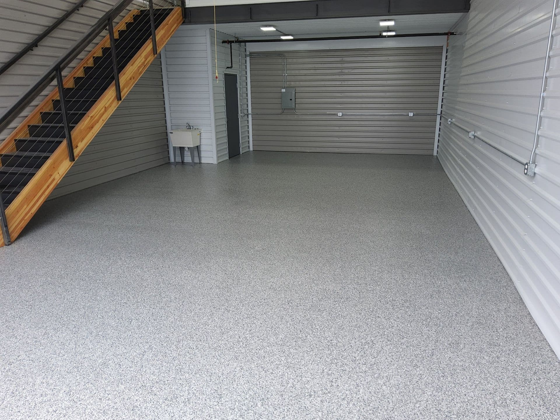 A storage unit with a closed garage door at the far end and a staircase on the left side leading up to a loft area. The floor has a fresh gray speckled polyurea coating.